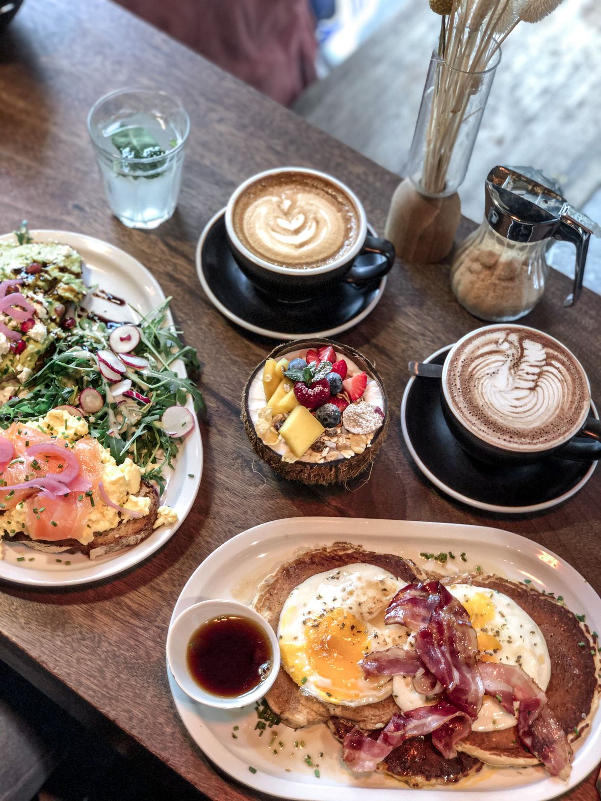 Image showing 2 plates with British breakfast on them, a desert in the middle, 2 cups of coffe, a glass of 
                            water, a sugar dispenser and a plant decoration, all on top of a brown table