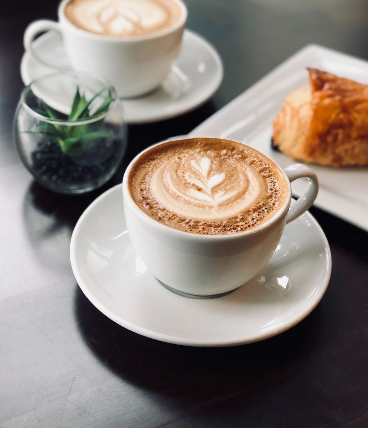 Image of a cup of coffee with a design on top with another cup of coffee, a croisant and 
                            a plant on the background all on a grey table