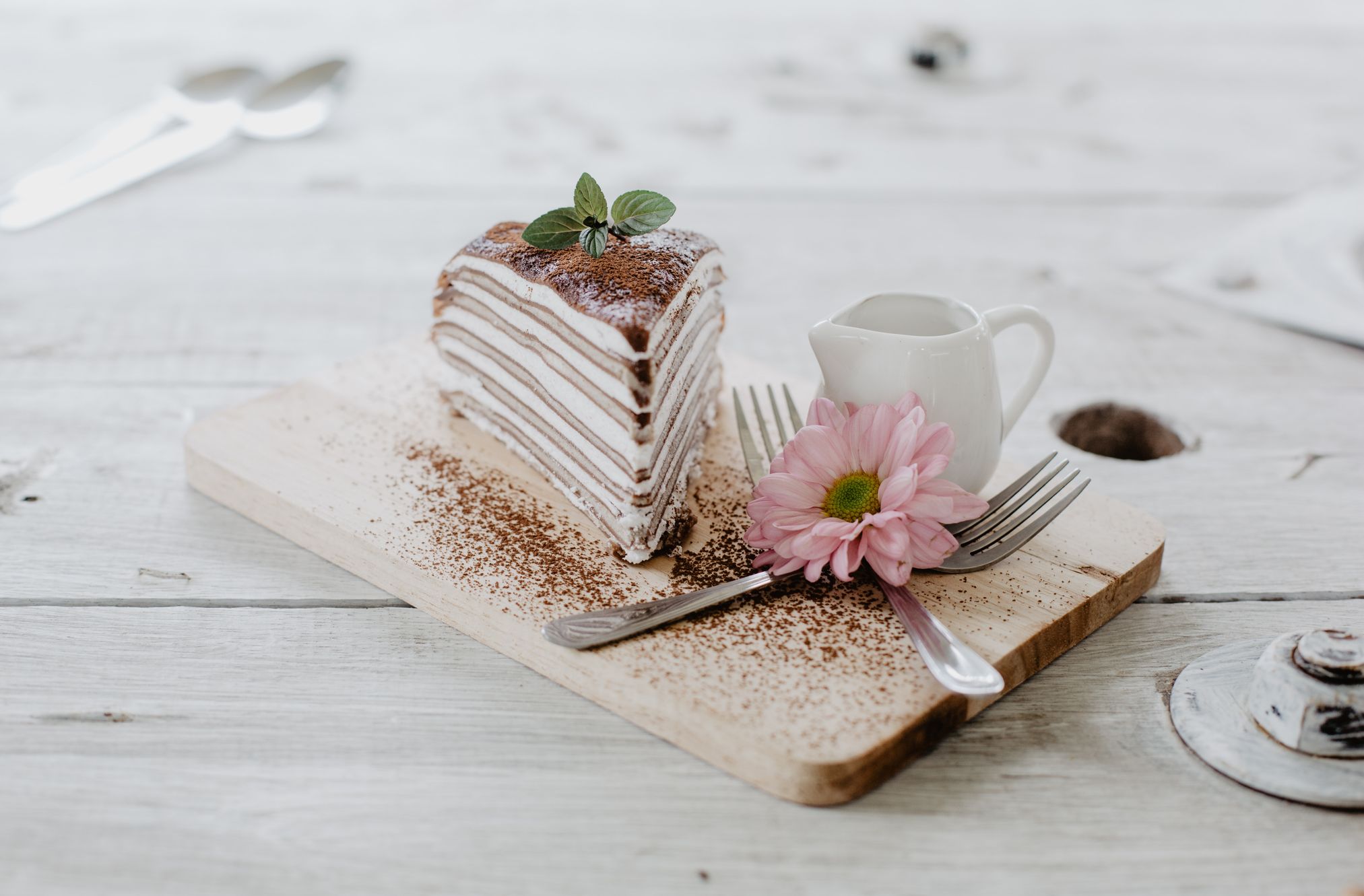 Picture of a white cake slice, utensils and a pink flower on a wooden board on top of a white table